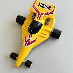 May include: Yellow toy racing car with black wheels and a red and blue stripe design. The car has a black windscreen and the words "FUSION" and "FOX" on the top. The car has the number "3" on the side.