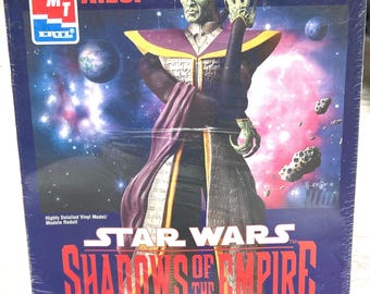 Puede incluir: Una caja de modelo para una figura de Star Wars Shadows of the Empire del personaje Kizor. La caja presenta un fondo morado y negro con una escena espacial y una figura de piel verde con una túnica morada. La caja está etiquetada como "Kizor" y "Star Wars Shadows of the Empire Collector Edition".