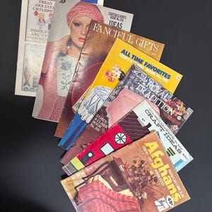 May include: A stack of vintage craft books with colourful covers featuring cross stitch, crochet, and afghan patterns. The titles include "More Sayings in Cross Stitch", "Fanciful Gifts", "All Time Favorites", "A Royal Tradition", "Craft Ideas", and "Afghans".
