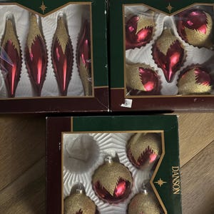 May include: Three boxes of Christmas baubles. Each box contains red and gold baubles. The boxes are green with the brand name "DANSON" printed on them. The baubles are in various shapes, including teardrops and round balls.