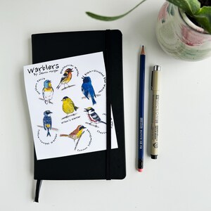 May include: A black notebook with a printed card featuring watercolour illustrations of various warblers. A blue pencil and a black pen are placed next to the notebook. The card includes the text "Warblers".