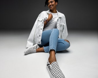 May include: A woman wearing a white denim jacket, blue jeans, and a pair of black and white patterned slip-on shoes. She is sitting on a white surface and looking to the left.