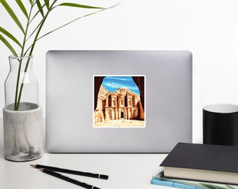 May include: A sticker featuring a view of the Treasury building in Petra, Jordan. The building is carved into the sandstone cliffs and has a facade with columns and arches. The sticker is a light brown color with a white border.