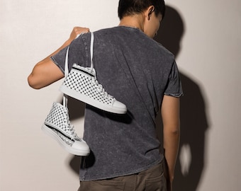 May include: White high-top sneakers with a black and white checkered pattern. The sneakers have a white sole and black accents. The shoes are being carried over a person's shoulder.