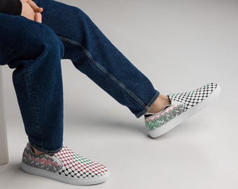 May include: Slip-on sneakers with a white sole and a patterned design. The pattern includes red, green, and black geometric shapes. The shoes are worn with blue jeans.