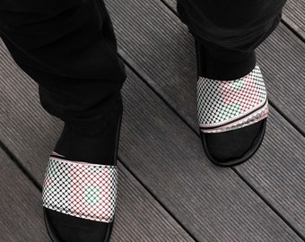 May include: Black slide sandals with a white, red, and green checkered pattern on the top strap. The sandals are worn with black pants and socks, and are set against a wooden deck.