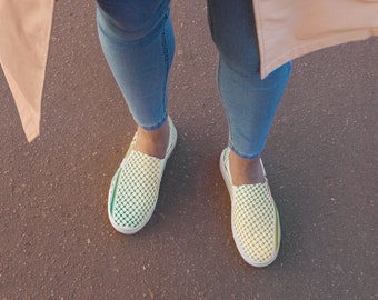May include: A pair of slip-on shoes with a white sole and a patterned upper. The pattern transitions from green to yellow dots. The shoes are worn with blue jeans and a tan coat.