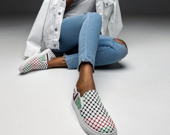 May include: White slip-on shoes with a black, red, and green geometric pattern. The shoes are worn by a person sitting on the floor with their legs crossed.