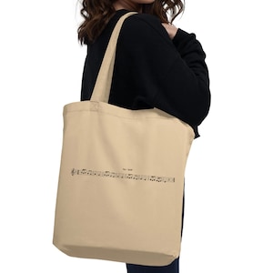 Op de afbeelding: Een beige tote bag met een zwart muzieknootontwerp en de tekst "Beat It" gedrukt op de voorkant.