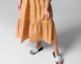 May include: A person wearing a long, tiered, orange dress and black slide sandals. The sandals have a white background with a blue eye pattern. The person is holding yellow sunglasses.
