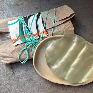 Op de afbeelding: Een helder, doorschijnend, golvend glazen object ligt op een beige keramisch bord. Het bord is ovaalvormig. Het glazen object is ingepakt in bruin papier en vastgebonden met een groen touwtje. Het papier heeft een label met de tekst "@artstrongs" erop.