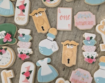May include: Assortment of Alice in Wonderland-themed cookies. Includes cookies shaped like a dress, clocks, teacups, a key, a door, and a bottle labeled "Drink Me." Some cookies have the text "one" and "Ariah in Wonderland."