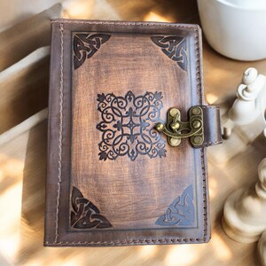 May include: A brown leather journal with Celtic knotwork corner designs and a central embossed pattern. The journal has a brass clasp closure and a stitched border. The cover has a distressed finish, giving it a vintage look.