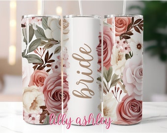 May include: A white tumbler with a floral design in shades of pink, beige, and green. The word "bride" is written in gold script on the tumbler.