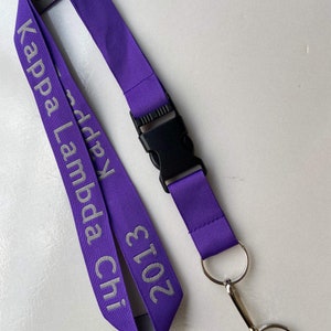 May include: Purple lanyard with a silver inscription that reads "Kappa Lambda Chi 2013". The lanyard has a black plastic clip and a silver key ring.