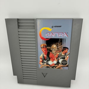 Pode incluir: Um cartucho de videogame cinza para Nintendo Entertainment System do jogo Contra. O rótulo apresenta o logotipo da Konami, o título Contra em letras grandes vermelhas e brancas e uma ilustração de dois soldados musculosos com armas.