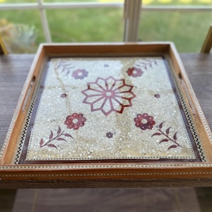 Puede incluir: Una bandeja de servir de madera con una incrustación decorativa de motivos florales blancos y rojos. La bandeja tiene un marco de madera marrón y una tapa de vidrio transparente.