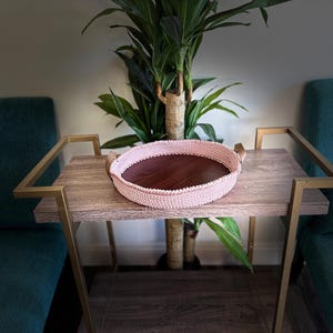 以下が含まれることがあります： 金色の金属製の脚が付いた木製のテーブルの上に、革の取っ手が付いたピンクの編み物トレイが置かれています。トレイは空で、木製の底があります。