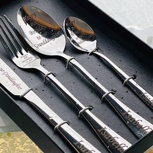 May include: A silver-coloured, engraved cutlery set presented in a black box. The set includes a knife, fork, and two spoons. The knife and fork are engraved with "Happy 4th Birthday, to our Granddaughter". The spoons are engraved with "Happy Birthday to Granddaughter" and the name "Isabella".