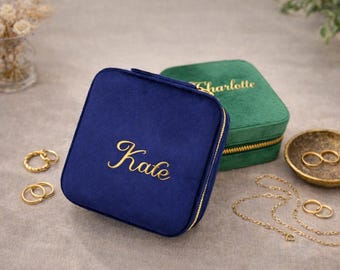 May include: A navy blue velvet jewelry case with the name "Kate" embroidered in gold script. A green velvet jewelry case with "Charlotte" embroidered in gold is behind it. Gold jewelry, including rings and a necklace, is displayed nearby.