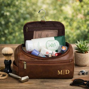 May include: A brown leather toiletry bag with the initials "MD" in gold. The bag is open, revealing various cosmetic products. A shaving brush, razor, and sunglasses are on the wooden surface.