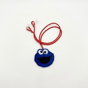 May include: A blue felt character face pendant with large white eyes and a wide black mouth, attached to a thin red cord with two clear plastic fasteners at the ends. The circular blue embroidery features a recognizable monster design.