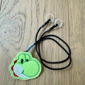 May include: A green Yoshi-themed lanyard with a black cord and white ear loops. The Yoshi character is embroidered with a light green body, red accents, and a white belly. The lanyard is designed to hold face masks or glasses.