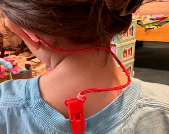 Puede incluir: Un cordón rojo con un pequeño clip de plástico rojo sujeto al cuello trasero de una camiseta azul claro. El cordón se extiende desde el clip hacia arriba en dirección al cuello, ofreciendo una forma segura de sujetar objetos a la prenda.