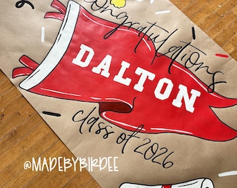 Könnte beinhalten: Ein Abschlussbanner aus braunem Kraftpapier mit einem roten Wimpel mit der Aufschrift "DALTON" und "Klasse 2026". Das Banner zeigt auch eine Doktorhut, eine Schriftrolle und das Wort "Glückwunsch".