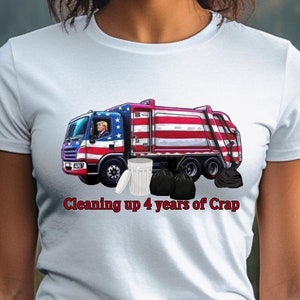 May include: A light blue t-shirt featuring a graphic of a garbage truck with an American flag design. The text "Cleaning up 4 years of Crap" is printed below the truck. The shirt is worn by a person.