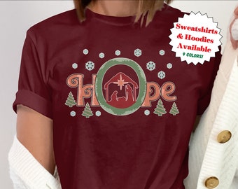 May include: A maroon t-shirt with a Christmas-themed design featuring the word "HOPE" in a retro font. The design includes a manger scene with a star and a wreath. The shirt is available in 9 colors.