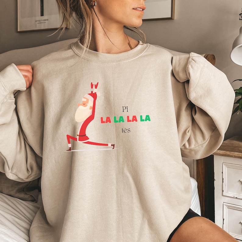 Puede incluir: Una sudadera beige con una imagen de dibujos animados de Pap&aacute; Noel haciendo yoga con el texto "Pi La La La La Les" impreso en la parte delantera.