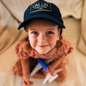 May include: A young girl wearing a blue baseball cap with the text "LOVE LIFE AT EVERY ALTITUDE" is sitting on a white couch and holding a toy airplane.