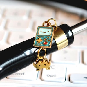 May include: A gold-colored charm with a retro video game design and a small gold-colored alien charm. The video game charm is teal with a smiling face and red accents. The charms are attached to a black object with a gold band.
