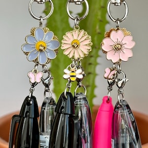 May include: Three pen charms with floral and insect designs. Each charm features a different flower, a butterfly, or a bee. The pens are black and pink, with silver hardware.