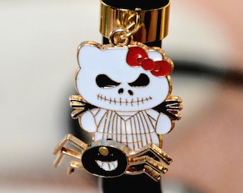 May include: A Halloween-themed charm featuring a white bear with a Jack Skellington-inspired face and a red bow. The charm includes a gold spider with a smiling face. The charm is attached to a black and gold metal object.