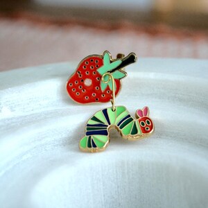 May include: A whimsical enamel pin set featuring a red strawberry and a green and blue caterpillar with a gold-tone finish. The strawberry has black seeds and a green stem. The caterpillar has pink ears and a red face.
