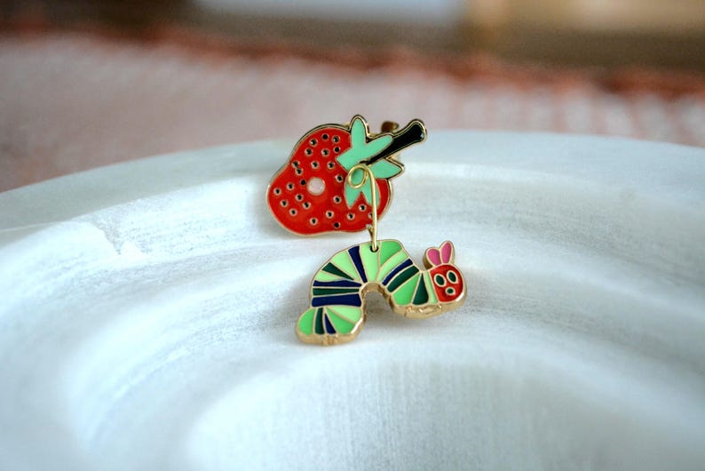 May include: A whimsical enamel pin set featuring a red strawberry and a green and blue caterpillar with a gold-tone finish. The strawberry has black seeds and a green stem. The caterpillar has pink ears and a red face.