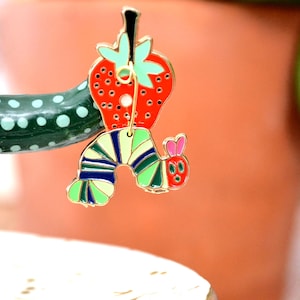May include: A colorful enamel pin featuring a red strawberry with green leaves and a whimsical caterpillar design. The caterpillar has a striped body in shades of green, blue, and yellow, with a red head and pink ears. The pin is attached to a green cactus.