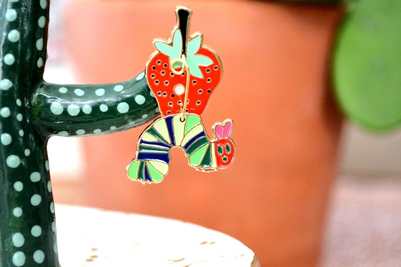 May include: A colorful enamel pin featuring a red strawberry with green leaves and a whimsical caterpillar design. The caterpillar has a striped body in shades of green, blue, and yellow, with a red head and pink ears. The pin is attached to a green cactus.