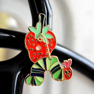 May include: A colorful enamel charm featuring a red strawberry with green leaves and a cartoon caterpillar. The caterpillar has green and blue stripes, a pink head, and is attached to the strawberry with a gold-colored wire.