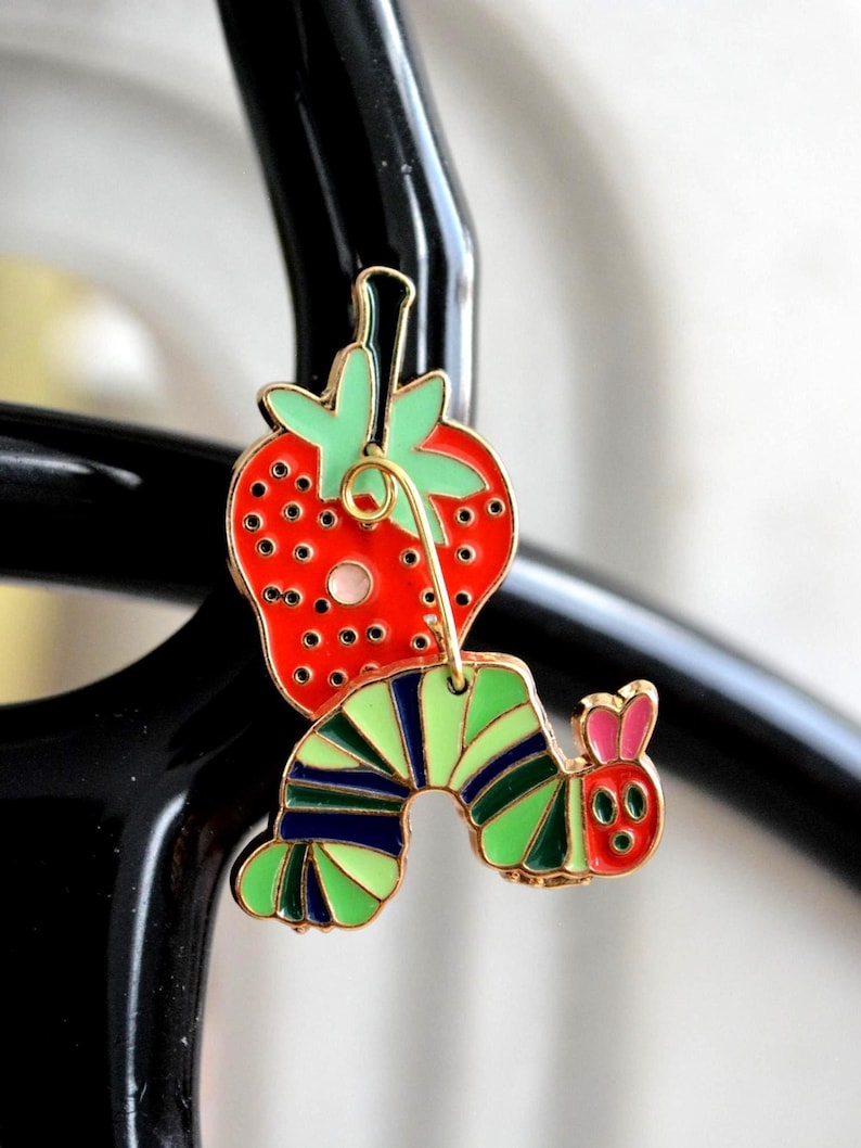 May include: A colorful enamel charm featuring a red strawberry with green leaves and a cartoon caterpillar. The caterpillar has green and blue stripes, a pink head, and is attached to the strawberry with a gold-colored wire.