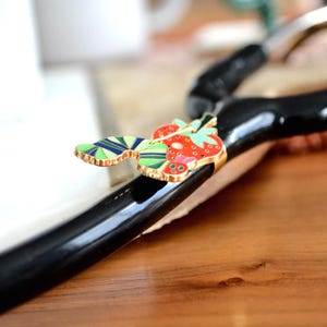 May include: A black, curved object with a gold-trimmed, colorful strawberry and leaf design. The strawberry is red with white seeds, and the leaves are green and blue. The object is on a wooden surface.