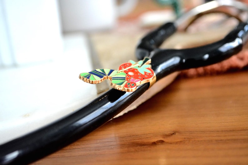 May include: A black, curved object with a gold-trimmed, colorful strawberry and leaf design. The strawberry is red with white seeds, and the leaves are green and blue. The object is on a wooden surface.