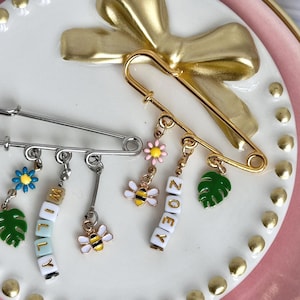 May include: Two safety pins with decorative charms. One silver pin has charms that include a blue flower, a green leaf, and the name "WILLY" spelled out in white letter beads. The other gold pin has charms with a bee, a pink flower, a green leaf, and the name "ZOEY".