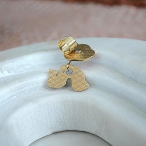 May include: A gold-colored earring with a unique, abstract design. The earring features a textured surface and a small, sparkling accent. The earring is set against a white, curved surface, creating a contrast in color and texture.