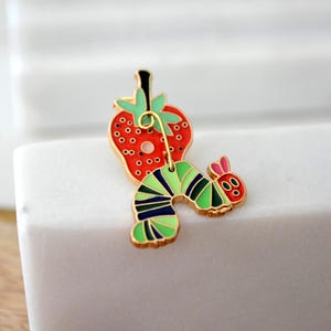May include: A colorful enamel pin featuring a red strawberry with black seeds and a green stem, connected to a green, black, and blue striped caterpillar with a red head and pink ears. The pin has a gold-colored border.