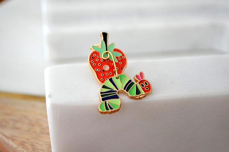 May include: A colorful enamel pin featuring a red strawberry with black seeds and a green stem, connected to a green, black, and blue striped caterpillar with a red head and pink ears. The pin has a gold-colored border.
