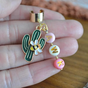 May include: Gold-tone ear cuff with a dangling charm featuring a green cactus with a bee and flower design, and pink and white beads spelling "AMK". The ear cuff is a unique piece of jewelry.