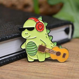 May include: A green dinosaur wearing red headphones and sunglasses is playing a yellow ukulele. The dinosaur is a enamel pin.